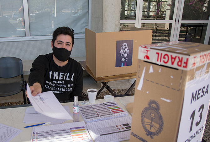 Un presidente de mesa con barbijo y sentado entrega una boleta de sufragio y hay una urna sobre la mesa junto a varias planillas y detrás está el biombo donde se puede votar