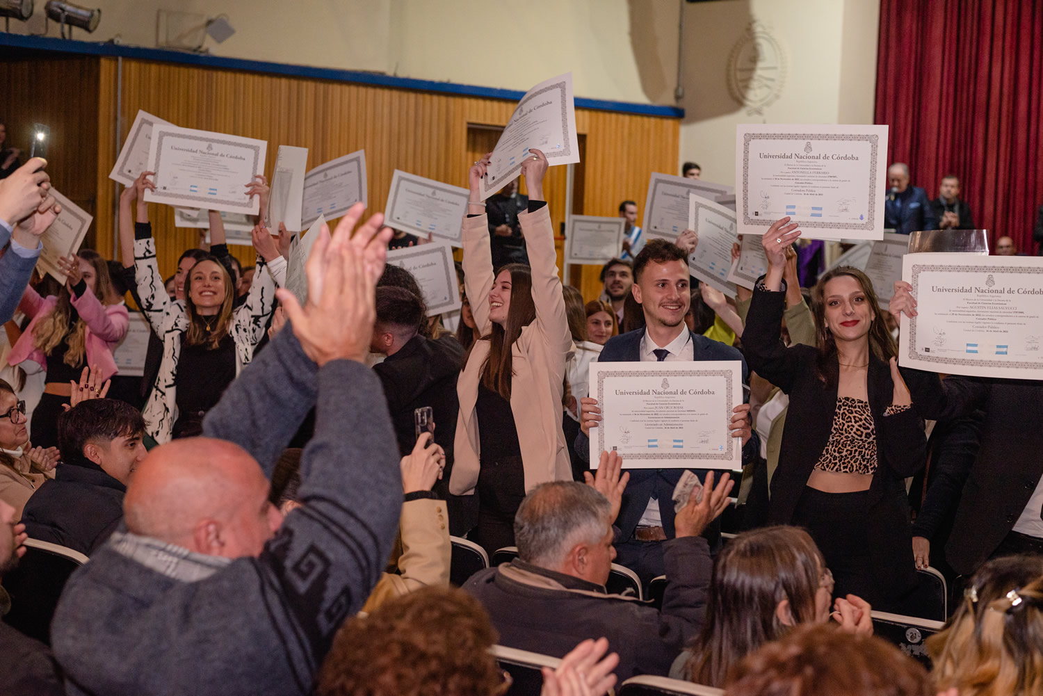 Un grupo de jóvenes que recibieron sus diplomas los elevan sobre sus manos ante la mirada de familiares y amistades en la Sala de las Américas del Pabellón Argentina