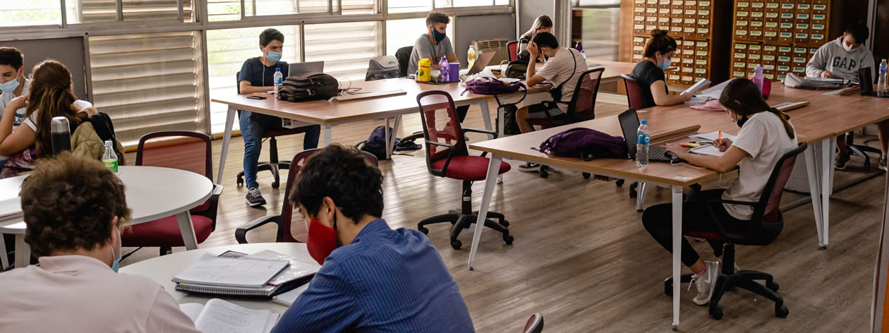 Una decena de estudiantes sentados en la Biblioteca usando barbijos leyendo y en los ventanales se advierte que es de día y hay sol