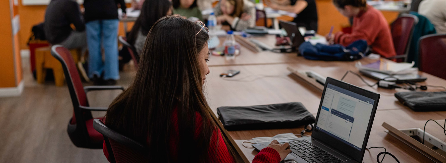 Una estudiante usando una computadora portátil en la biblioteca, sentada, y hay una docena de personas atrás sentados