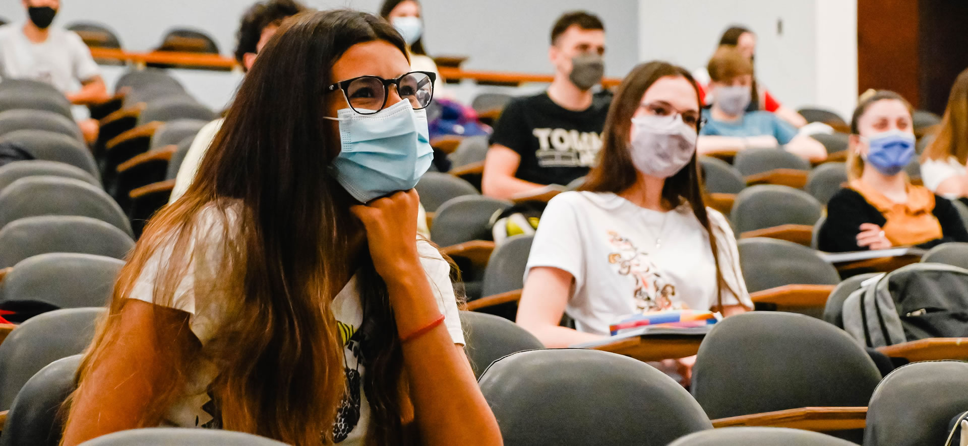 Una docena de estudiantes con barbijos sentados en una de las aulas de la Facultad, y una de las estudiantes está más cerca, con cabello largo y anteojos, apoyando su brazo y su puño en el mentón y sonriendo