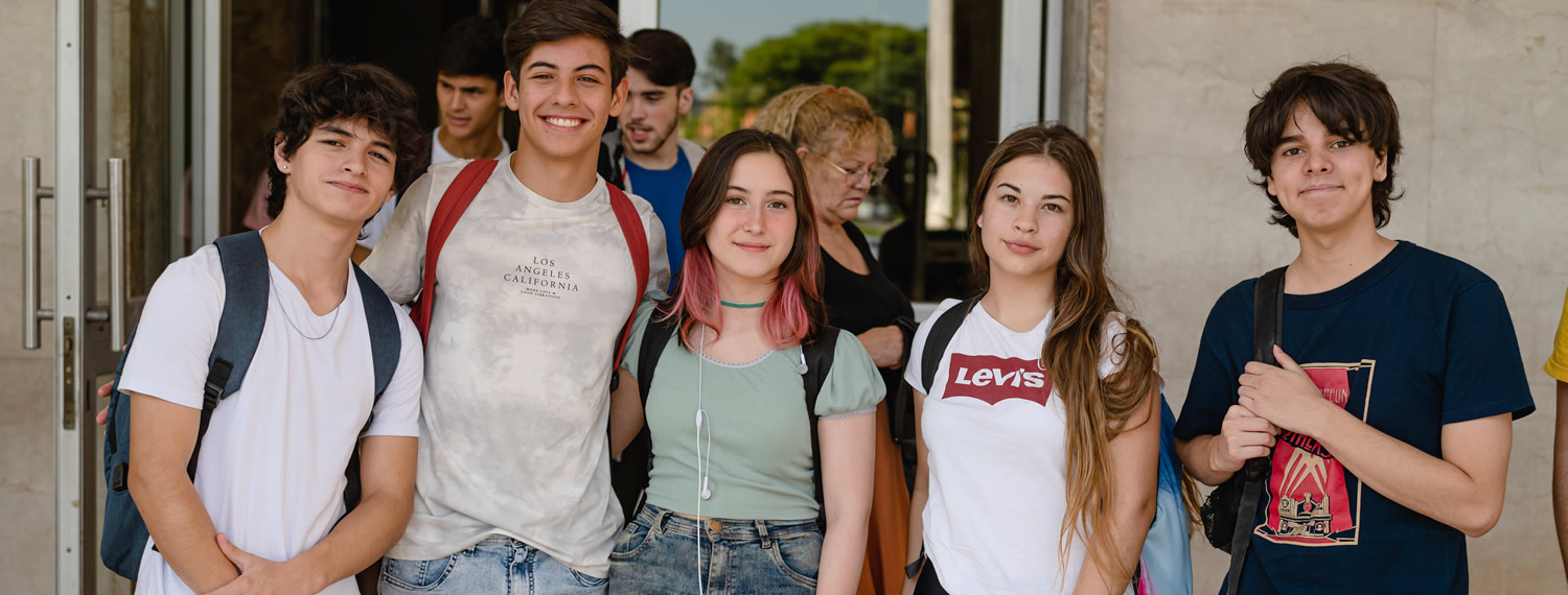 Tres estudiantes varones y dos estudiantes mujeres de pie sonriendo y detrás hay cuatro personas caminando frente al ingreso al Pabellón Argentina