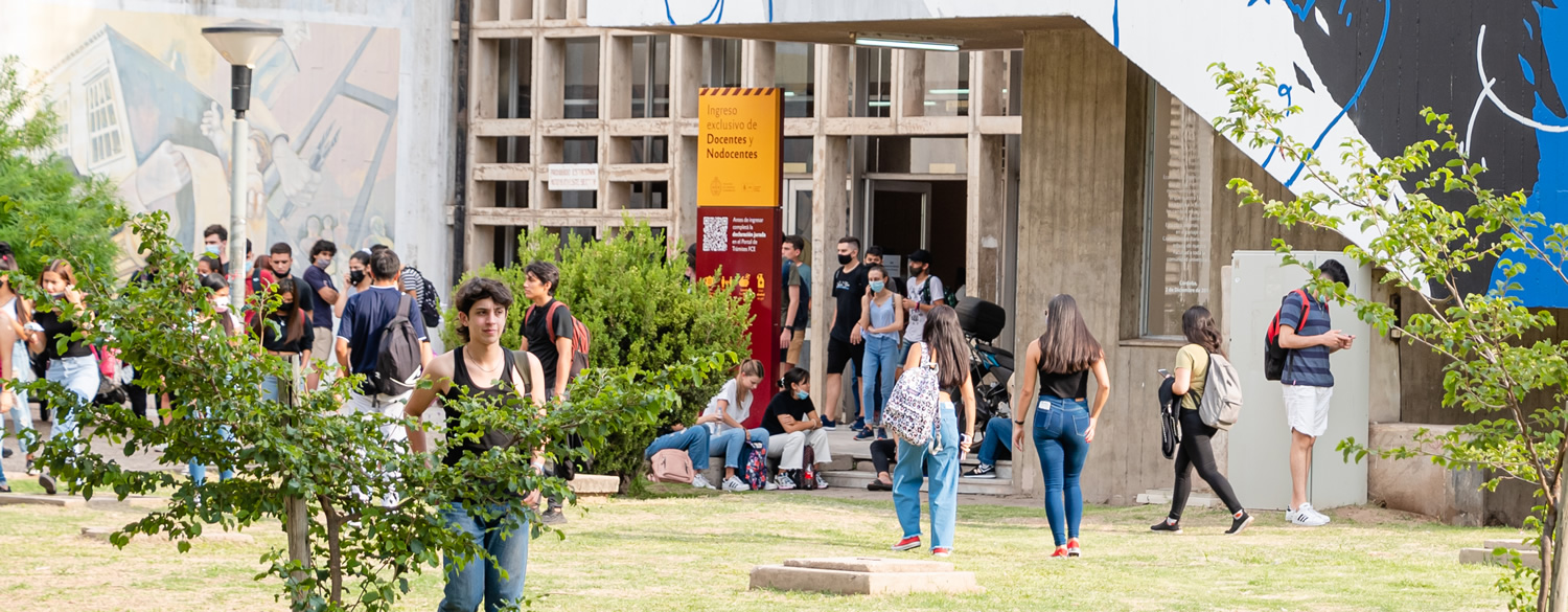 Una treintena de estudiantes en la puerta del acceso norte de la Facultad en un día soleado con pasto crecido y dos arbustos con hojas