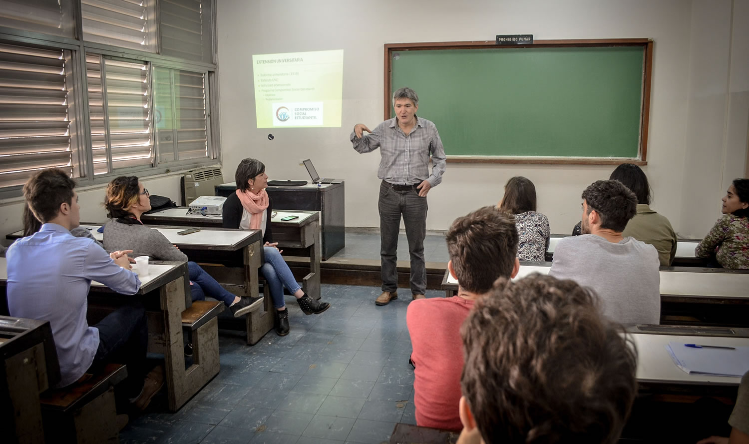 Imagen del profesor Juan Vega de pie explicando de qué trata el proyecto Güemes Solidario frente a una decena de personas en una aula de la Facultad