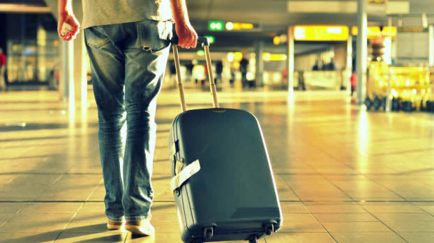 Imagen de un hombre de espaldas arrastrando una valija azul en un aeropuerto