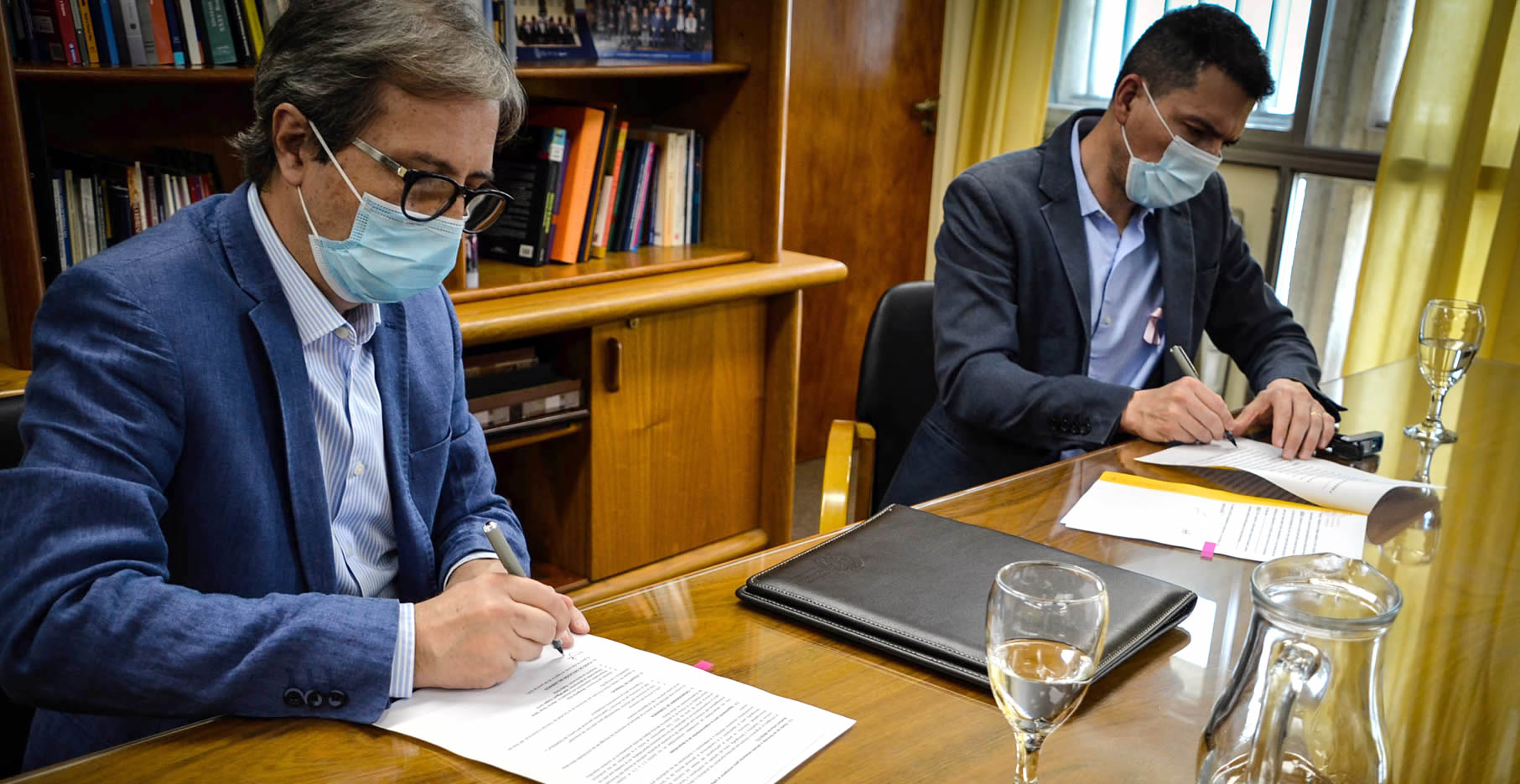 Jhon Boretto y Luis Picat, con barbijos, sentados firmando el convenio