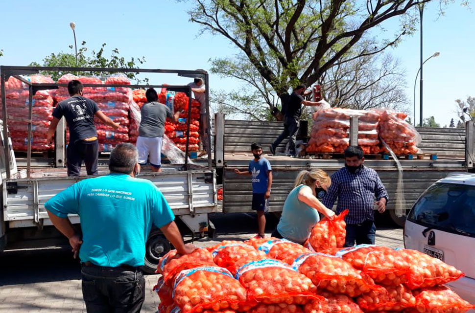 Tres varones llenan el trailer de un camión con bolsas de cebollas mientras un señor mira apoyado en varios lotes de bolsas de cebollas que son vistos por una señora y un señor