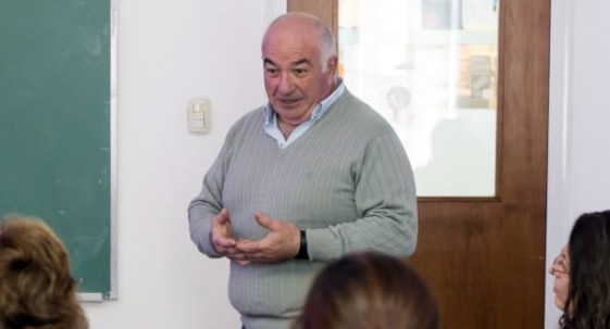 El profesor Beltramino explica en una clase. Se encuentra de pie con las manos entrelazadas y viste un pulover color gris