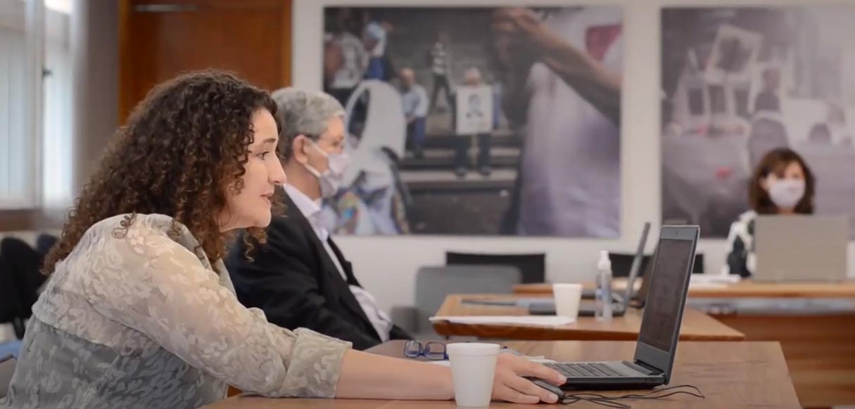 En una sala, de día, con paredes con dos grandes cuadros, la doctora Funes sentada hablando frente a una computadora portátil. Detrás está el doctor Zanazzi sentado, con barbijo, frente a una computadora portátil y a tres metros también sentada, con barbijo, está la vicedecana Catalina Alberto mirando su computadora portátil