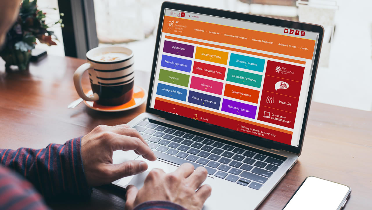 Una persona frente a una computadora portátil con la página web nueva de la Secretaría de Extensión y una taza de café al lado