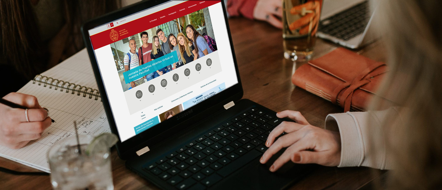 Una mujer mirando la nueva página web en su notebook sobre una mesa frente a una persona que escribe en un cuaderno