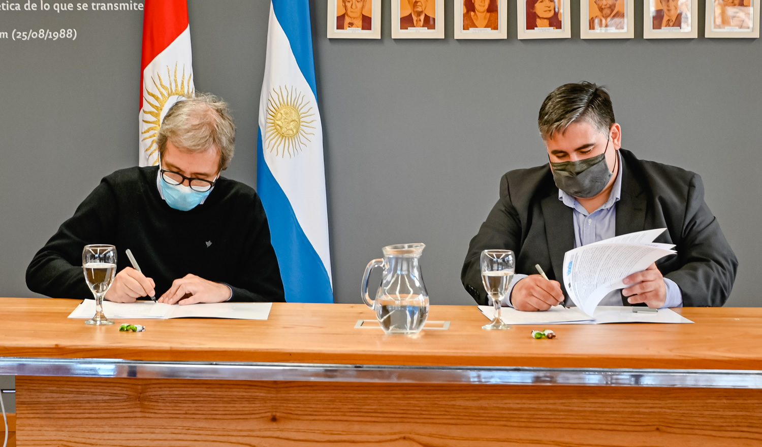 Jhon Boretto y Enzo Cravero, sentados y con barbijo, firmando copias del convenio específico