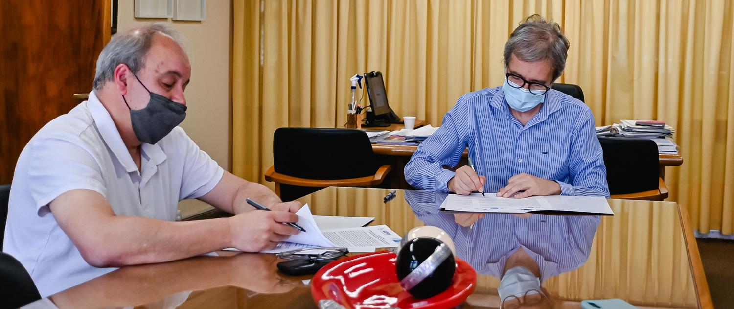 Javier Varetto y Jhon Boretto sentados en una mesa con cubierta de vidrio. Están en una oficina amplia con cortinas grandes en los ventanales, usando barbijos y firman los papeles del convenio.