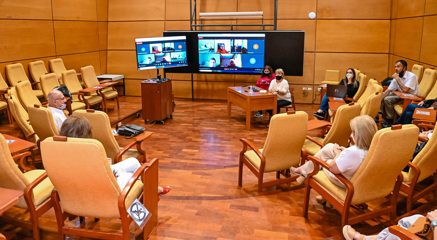Un auditorio de Graduados con dos pantallas gigantes encendidas conectadas a una sesión de Zoom y están sentadas diez personas en sillones grandes