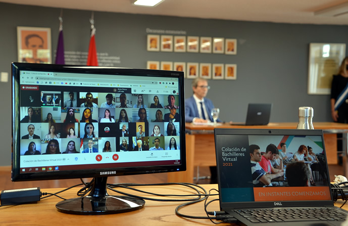 El decano Jhon Boretto sentado con saco y corbata frente a una computadora portátil. Más adelante, sobre otra mesa, un monitor con una sesión de Google Meet con cuarenta egresados y egresadas y una computadora portátil comandando la transmisión virtual