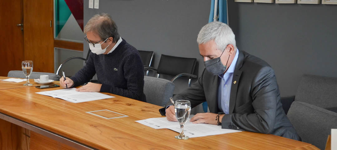 Jhon Boretto y Marcelo Jaluf, con barbijos, sentados firmando papeles sobre una mesa de la sala Camilo Dagum