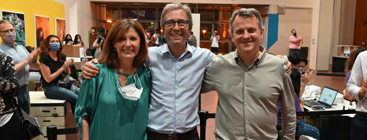 Catalina Alberto a la izquierda, Jhon Boretto al medio y Ricardo Decalzi a la derecha sonriendo, abrazados y hay una decena de personas aplaudiendo alrededor