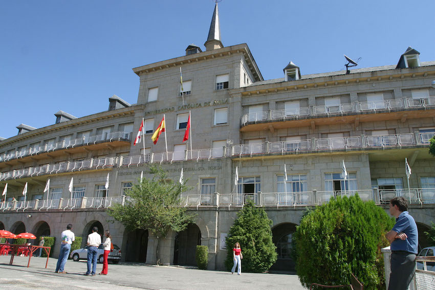 El edificio de la Universidad Católica de Ávila en un día soleado con tres banderas flameando sobre la puerta de entrada y hay cinco personas caminando sobre la explanada de acceso