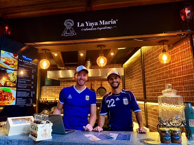 Luis Royo y su pareja, vistiendo camisetas de la selección argentina de fútbol y atendiendo su local gastronómico llamado La Yaya María detrás del mostrador
