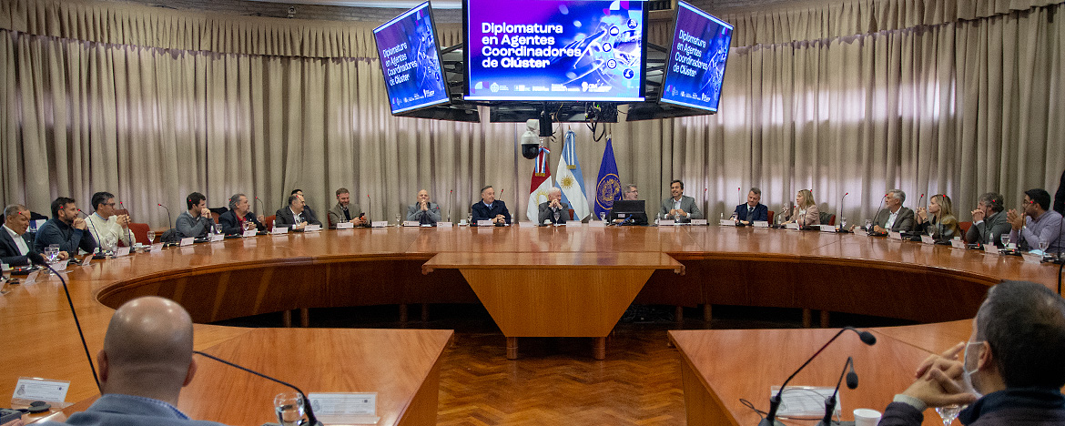 La sala del Consejo Superior con treinta personas sentadas y hay tres grandes televisores LED en el techo donde dice Diplomatura Coordinadores de Clusters y la sala tiene amplias cortinas en sus ventanales