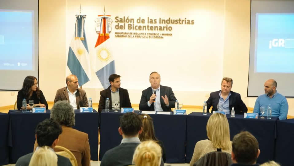 El ministro Acastello, sentado y con un micrófono hablando, acompañado por el doctor Ricardo Descalzi a su izquierda y cuatro autoridades más, y detrás hay una bandera argentina y otra de la Provincia de Córdoba, y frente a ellos, también sentados, hay siete personas