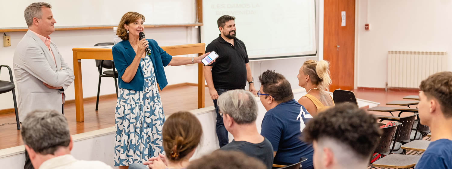 La decana Catalina Alberto habla de pie con un micrófono y a su derecha está el Vicedecano Ricardo Descalzi y a la izquierda el secretario Lautaro Blatto, mientras hay ocho personas sentadas en el aula