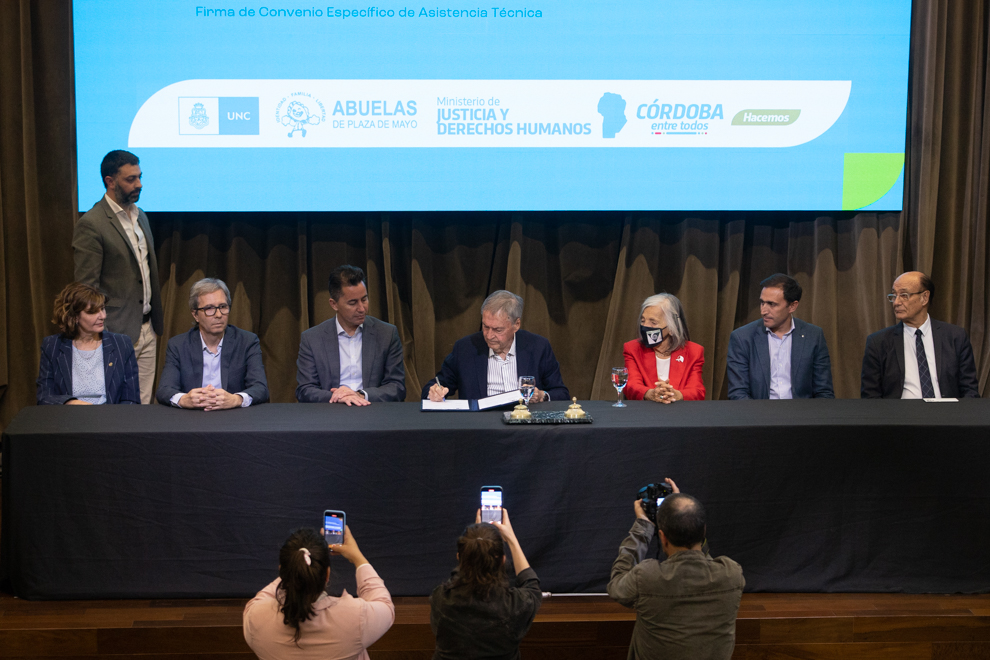 Autoridades provinciales y universitarias sentadas en una mesa larga con mantel y detrás hay una pantalla gigante con los logos de cada institución, mientras que adelante y debajo del estrado hay tres personas con cámaras de fotos