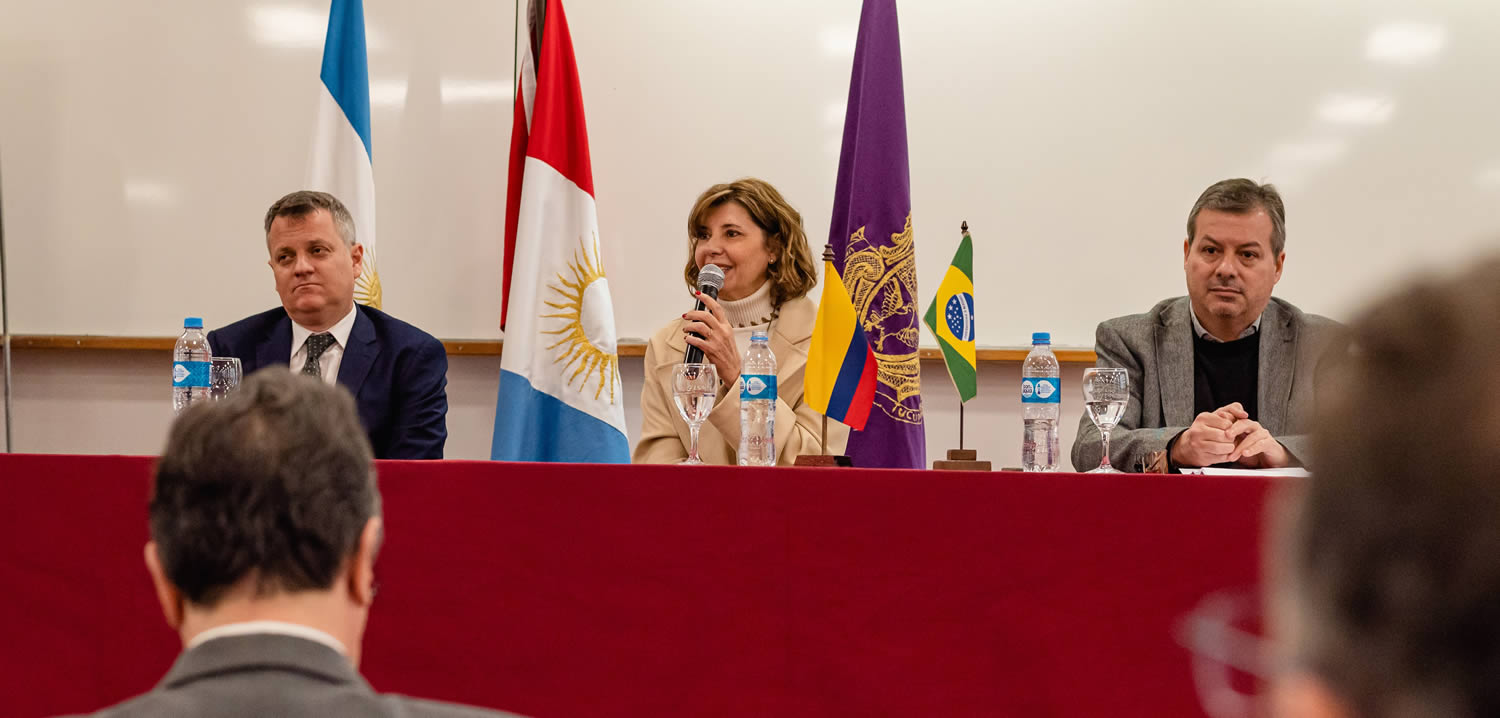 Ricardo Descalzi, Catalina Alberto hablando con un micrófono y Marcelo Capello, sentados, hay una mesa con mantel de paño grueso, hay tres botellas de agua mineral y tres copas, una banderita de Brasil y otra de Colombia, y detrás hay tres mástiles con banderas de Argentina, Provincia de Córdoba y la Universidad Nacional de Córdoba