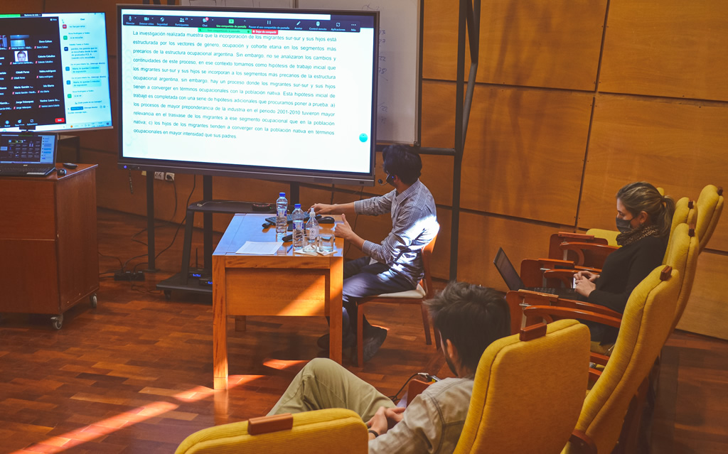 Un docente sentado frente a dos pantallas gigantes manejando un mouse y un teclado sobre una mesa de madera que tiene una botella y una copa con agua, y detrás están sentados una mujer con barbijo y un varón en un auditorio de la Escuela de Graduados con pisos y paredes de madera