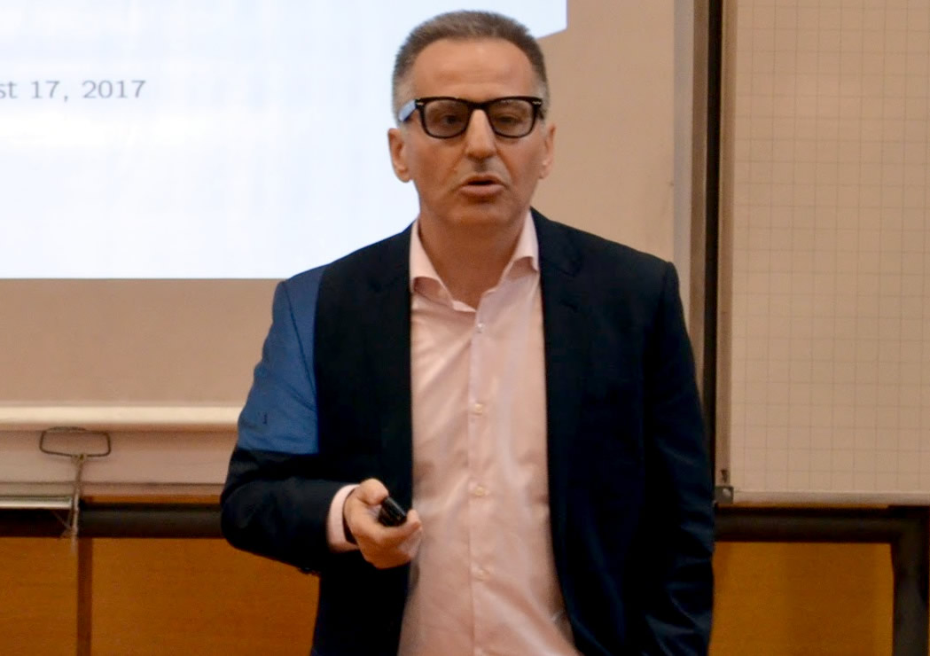 Martín Uribe usando lentes exponiendo de pie con un señalador en la mano usando saco y camisa y detrás hay un pizarrón de acrílico donde se proyecta una filmina