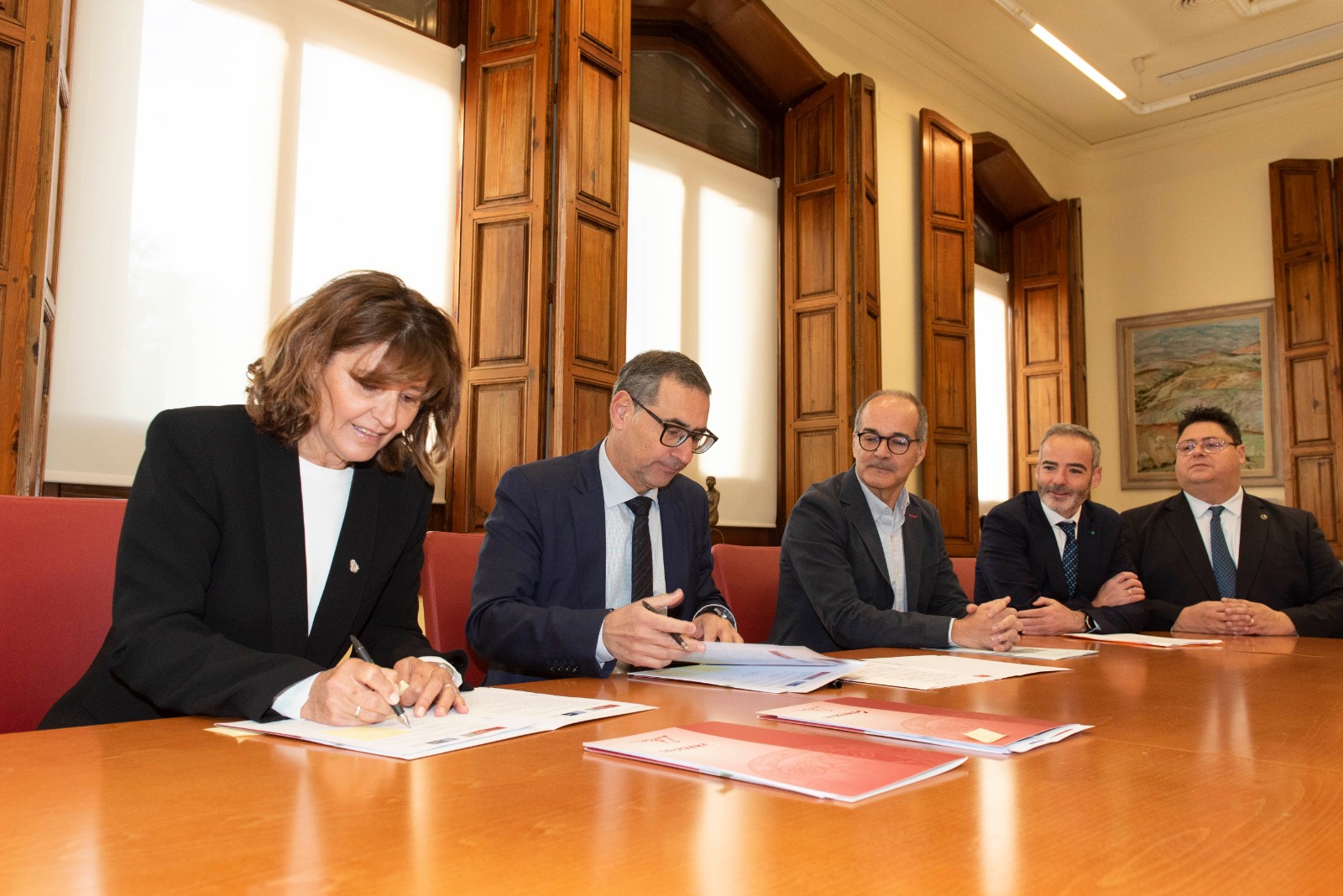 La Decana Catalina Alberto firma un documento sentada ante la mirada de cuatro funcionarios españoles también sentados en una mesa de madera dentro de un recinto con columnas de madera