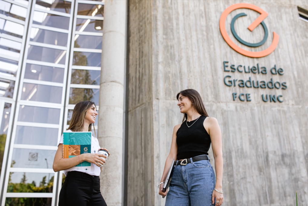 Dos estudiantes mujeres en el frente del edificio de la Escuela de Graduados, de pie, mirándose, sonrientes, y una de ellas tiene una carpeta en su brazo derecho