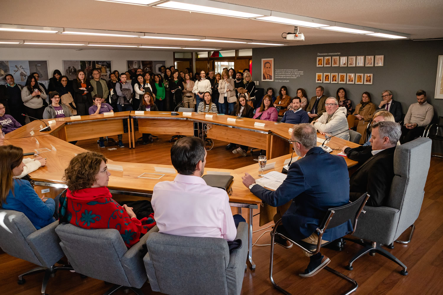 La sala del Consejo Directivo en un día luminoso colmada de personas mientras los consejeros, el Decano y el Vicedecano se encuentran sentados llevando a cabo una sesión plenaria