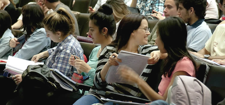 Cinco estudiantes mujeres sentadas con cuadernos en sus manos y una de ellas mira sonriente hacia atrás