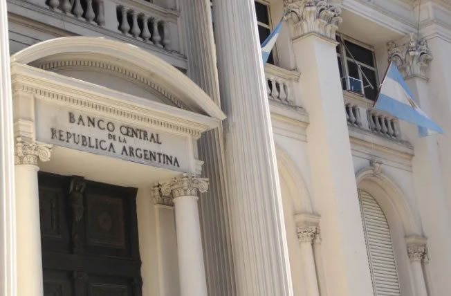 El frento del edificio del Banco Central, con dos banderas argentinas flameando, hay cinco columnas de estilo corintio y una puerta tallada de madera oscura, en un día luminoso