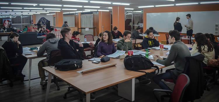 El espacio central de la Biblioteca con tres largas mesas con veinte estudiantes sentados y dos están de pie frente a pizarras