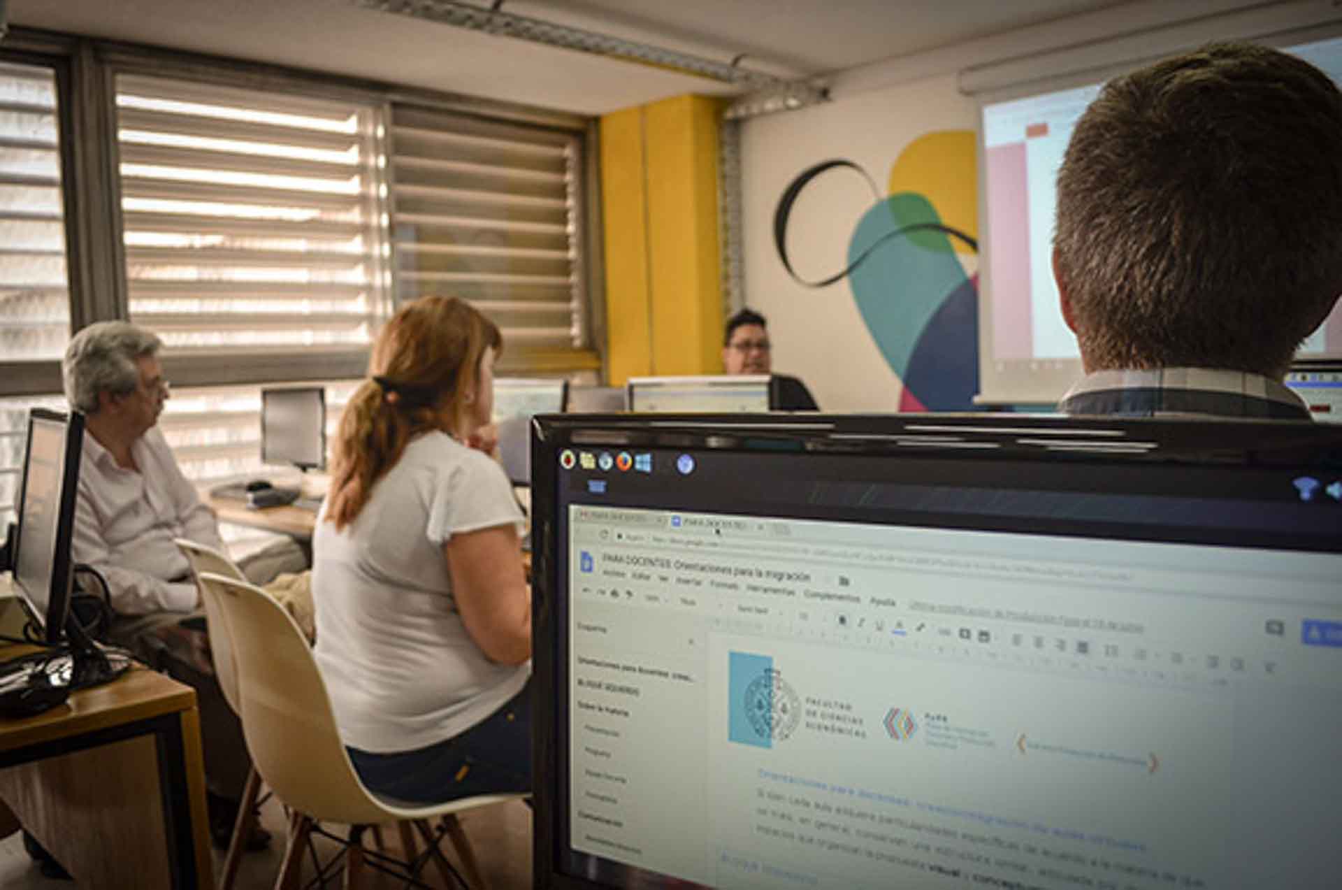 Tres docentes sentados frente a computadoras, dentro de un aula que tiene un mural pintado en una pared y un gran ventanal, observan la exposición de un docente que está de pie, en un día luminoso