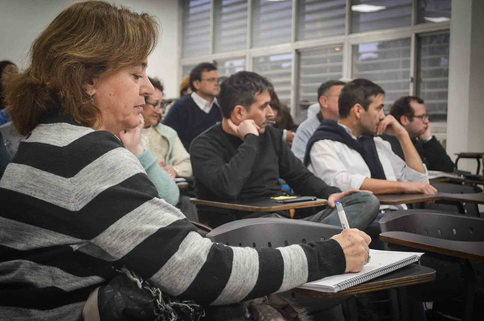 Una docente escribe sentada sobre un cuaderno y una decena de profesores sentados miran hacia el frente