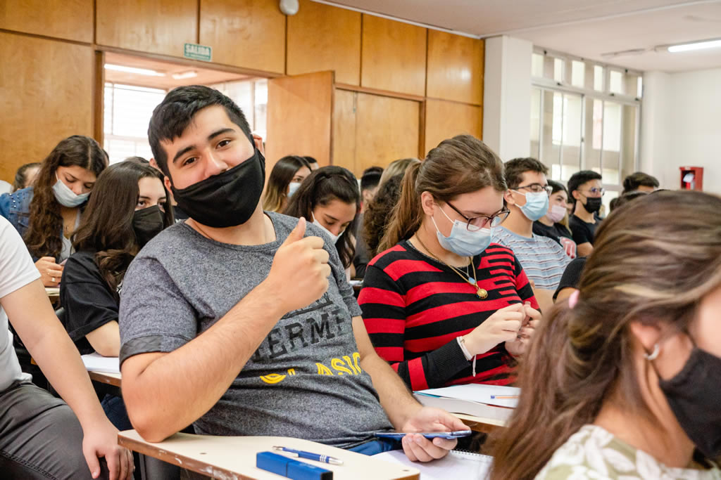 Un estudiante varón sentado con barbijo muestra su pulgar arriba y hay una treintena de estudiantes mujeres y varones sentados con barbijos