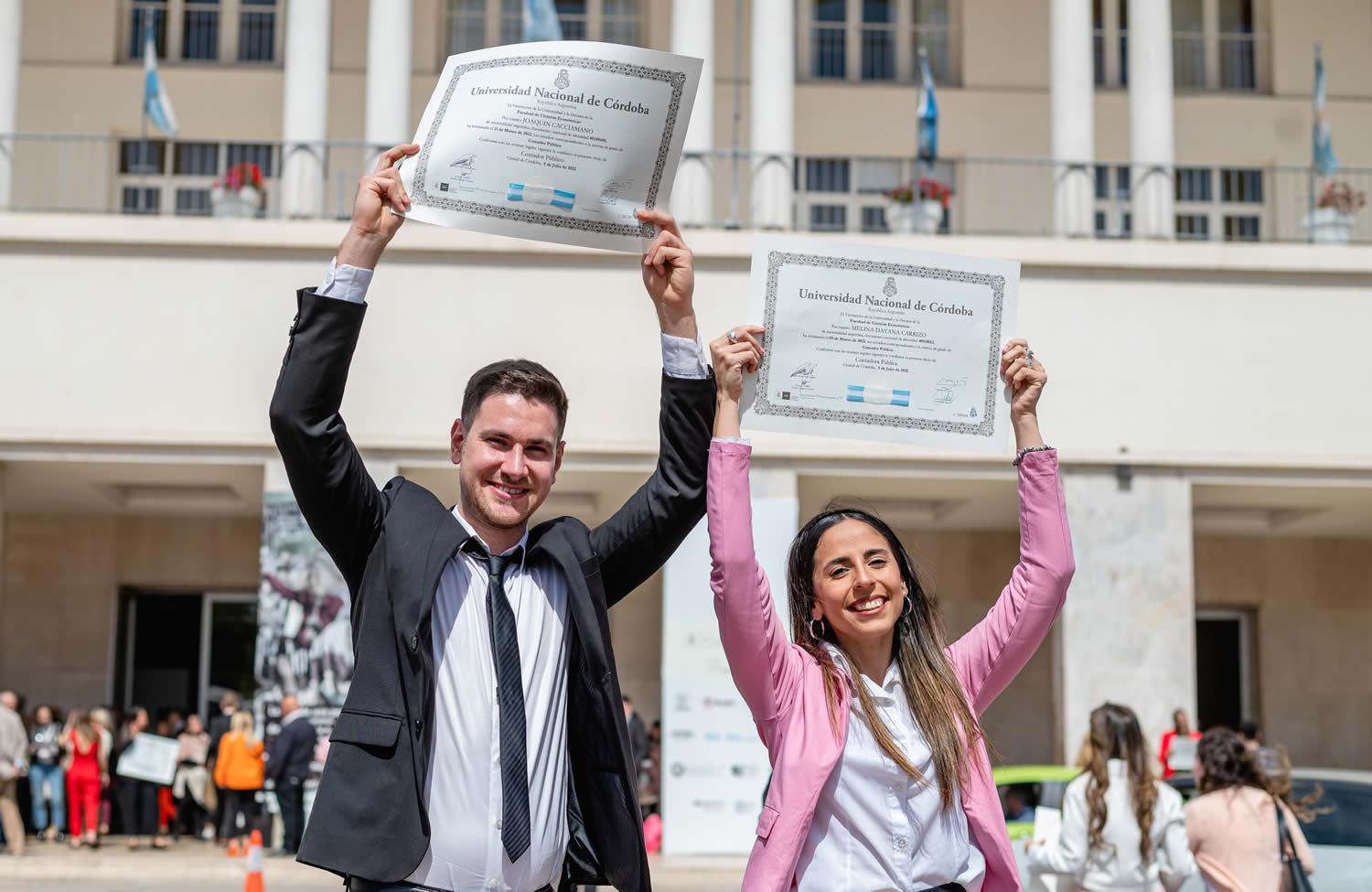 Un egresado con saco y corbata y una egresada con saco y cabello largo sonríen y levantan sus diplomas frente al Pabellón Argentina, al aire libre y en un día soleado al mediodía