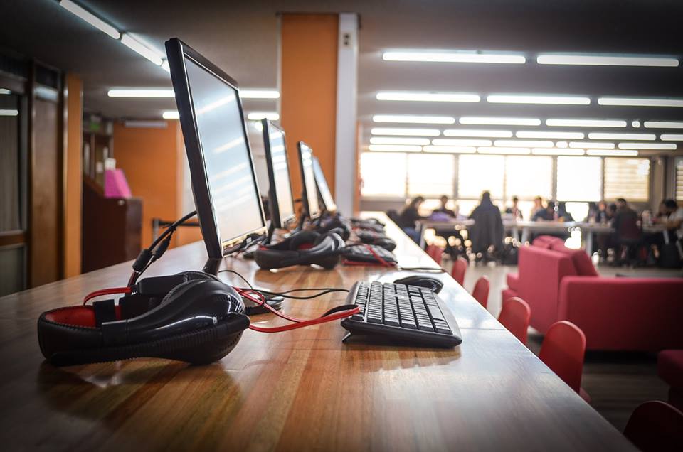 Una mesa alta con cuatro monitores, cuatro teclados de computadora, unos auriculares, mouses y al fondo están sentadas diez personas al lado de una ventana