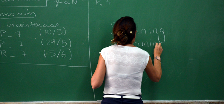 Una docente con blusa musculosa y con cabello recogido escribe en una pizarra con una tiza