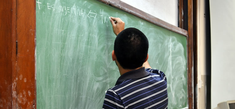 Imagen de un docente joven de espaldas escribiendo con una tiza blanca sobre un pizarrón verde
