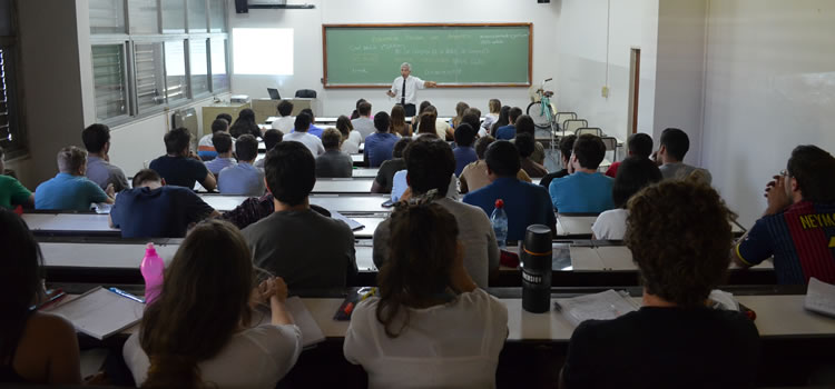 Una clase con una treintena de estudiantes sentados y un docente de pie explicando un tema en un día luminoso