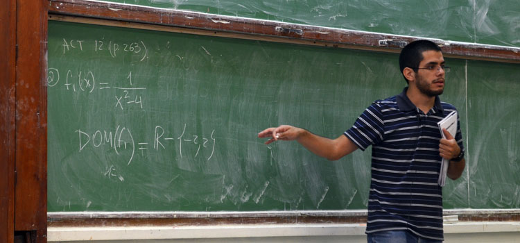 Un docente señalando la pizarra con un cuaderno en la mano