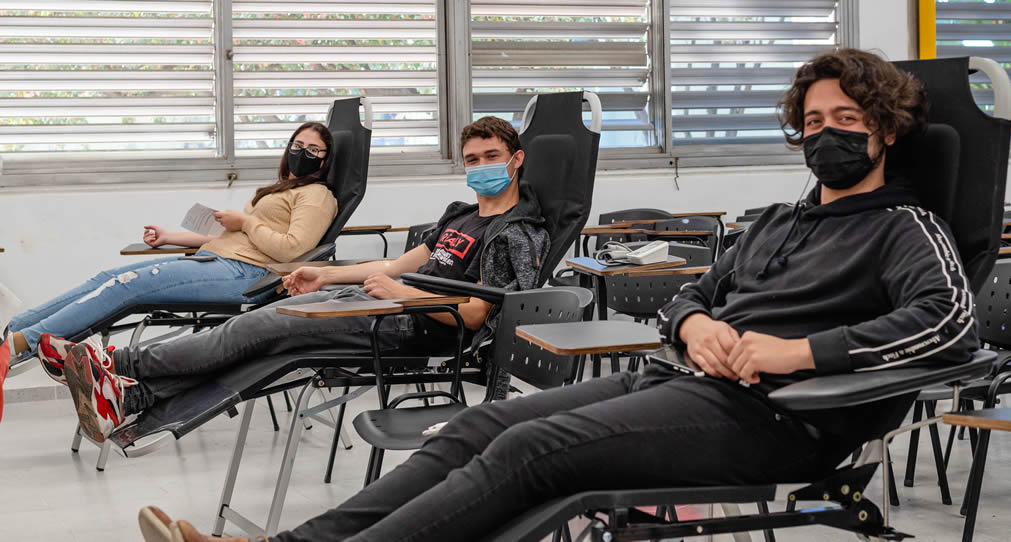 Dos chicos y una chica con barbijos en reposeras durante una donación de sangre, con sus brazos extendidos, en una aula grande de Económicas