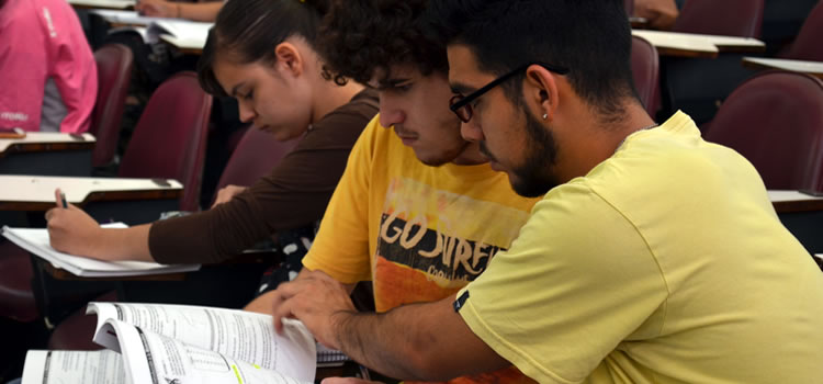 Un estudiante usando anteojos de lectura repasa un libro de texto junto a otro estudiante, ambos sentados, y detrás hay una estudiante mujer sentada escribiendo en un cuaderno con su mano izquierda