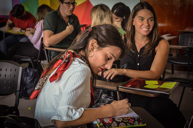 Una estudiante sentada escribe un cuaderno con una lapicera mientras otra estudiante sentada sonríe frente a ella y detrás hay seis estudiantes sentados escribiendo