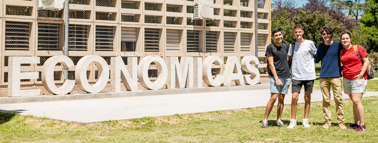 Tres estudiantes varones y una estudiante mujer parados juntos en el jardín de la Facultad sonriendo junto a las letras que dicen Económicas en un mediodía muy soleado