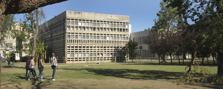 El edificio de la Facultad al fondo, con el parque de Ciudad Universitaria en un día soleado con tres estudiantes caminando